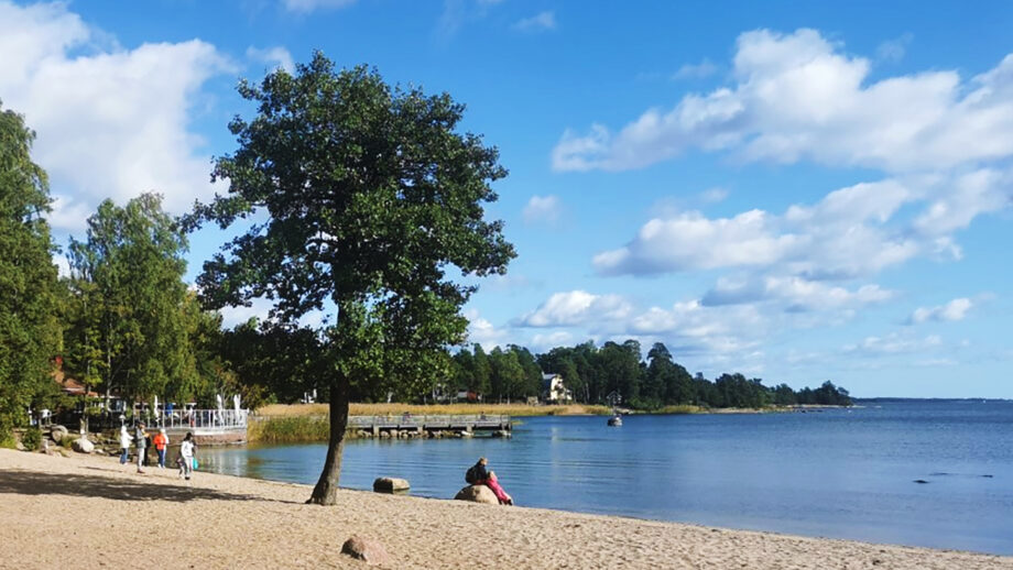 Kuvassa on kesäinen Matinkylänranta. Rannalla on hiekkaa, lehteviä puita, vesi on sininen. Taivas poutapilvinen.