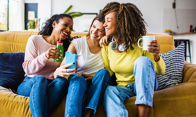 Kuvassa on kolme iloista nuorta naista viettämässä aikaa yhdessä sohvalla. Yhdellä on kädessään smoothie ja toisella kahvimuki. Keskimmäinen ottaa kolmikosta selfietä.