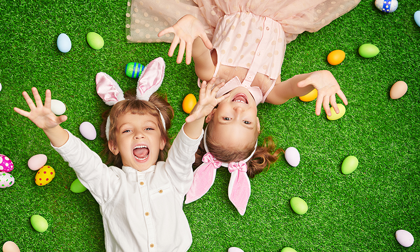 Kaksi iloista on selällään nurmikolla. Lapset katsoo ylöspäin kameraa kohden ja kurottelevat käsiään. Kummallakin lapsella on pupun korvat, heidän ympärillään, nurmikolla on värikkäitä pääsiäismunia.