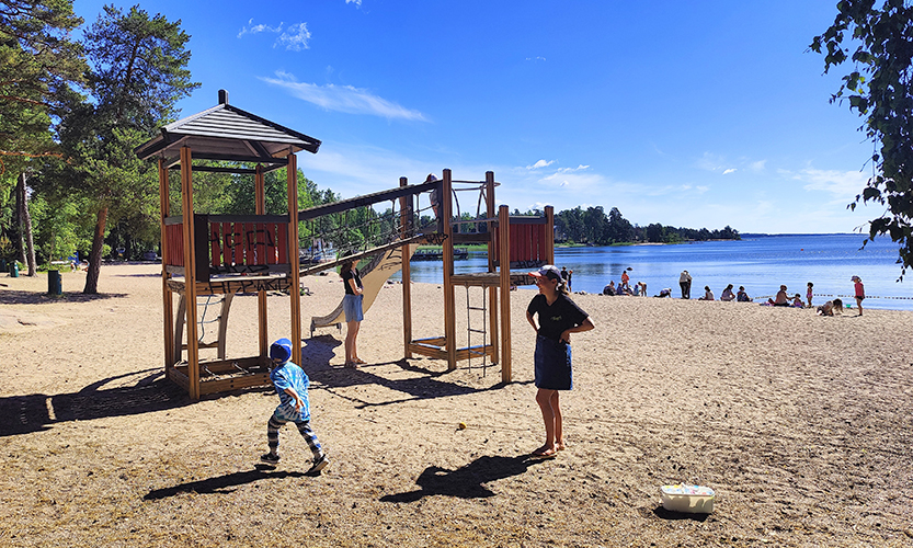 Aurinkoinen päivä meren rannalla. Kuvassa on etualalla pieni lapsi ja aikuinen sekä kiipeilyteline. Rannalla, vesirajassa on eri-ikäisiä ihmisiä.