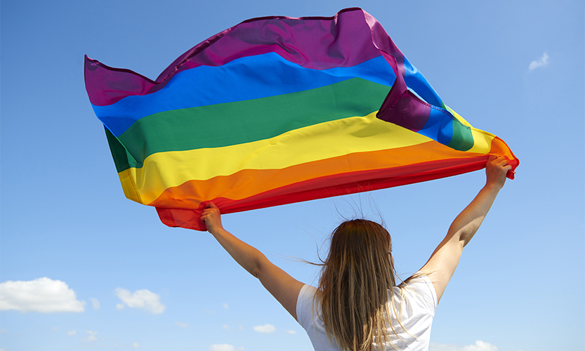 Kuvassa henkilö pitää korkealla päänsä päällä liehuvaa sateenkaarilippua.