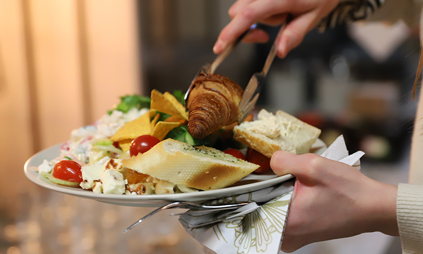 Kuvassa henkilö pitää käsissään ruokalautasta, jossa on salaattia, tomaattia, croissantteja ja monenlaisia ruokia.
